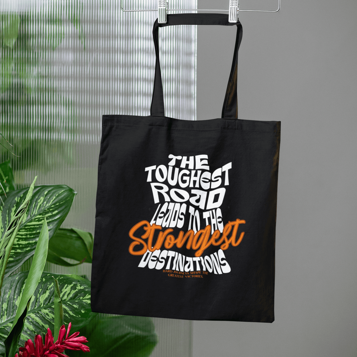 Eco tote bag with “The Toughest Road Leads to the Strongest Destinations” motivational typography design in white and orange on black organic cotton by LostLogicCo Canada.